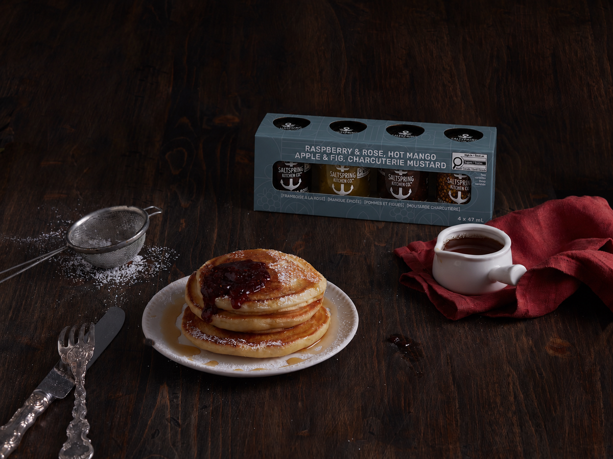 SaltSpring Kitchen Company Everyday Sampler Collection on a dark table top with a stack of pancakes with Raspberry and Rose Spread on them, with cutlery and syrup on the table with a napkin and icing sugar sifter