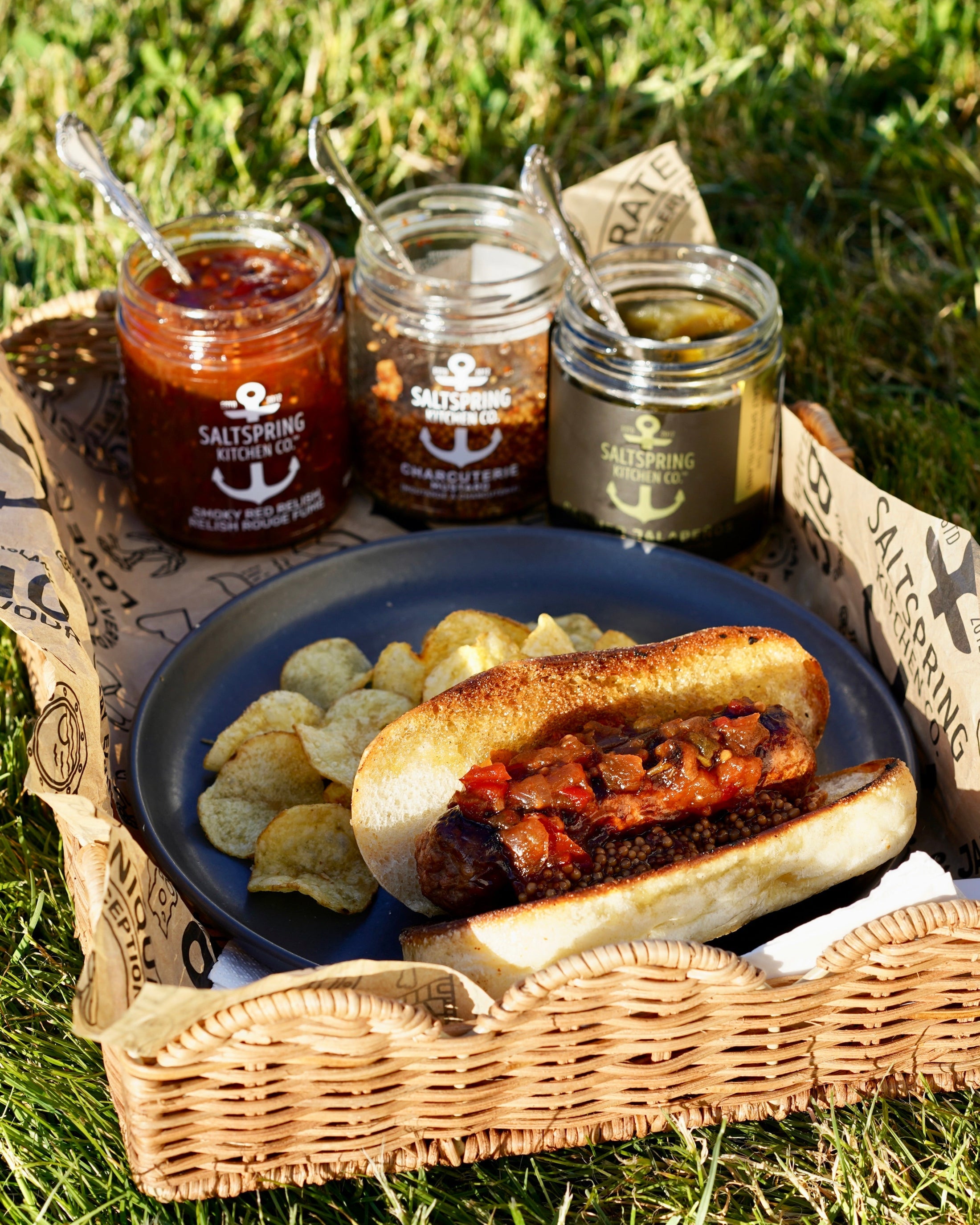 Vegan brat with SaltSpring Kitchen Company Smoky Red Relish and Charcuterie Mustard on a plate with potato chips and three jars of condiments in a picnic setting.