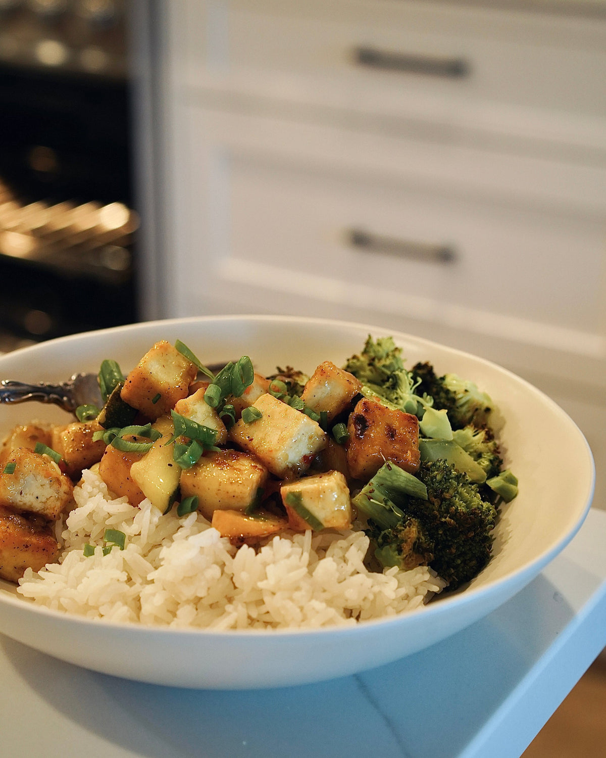 Bowl of food with rice, tofu, and broccoli on a kitchen counter.