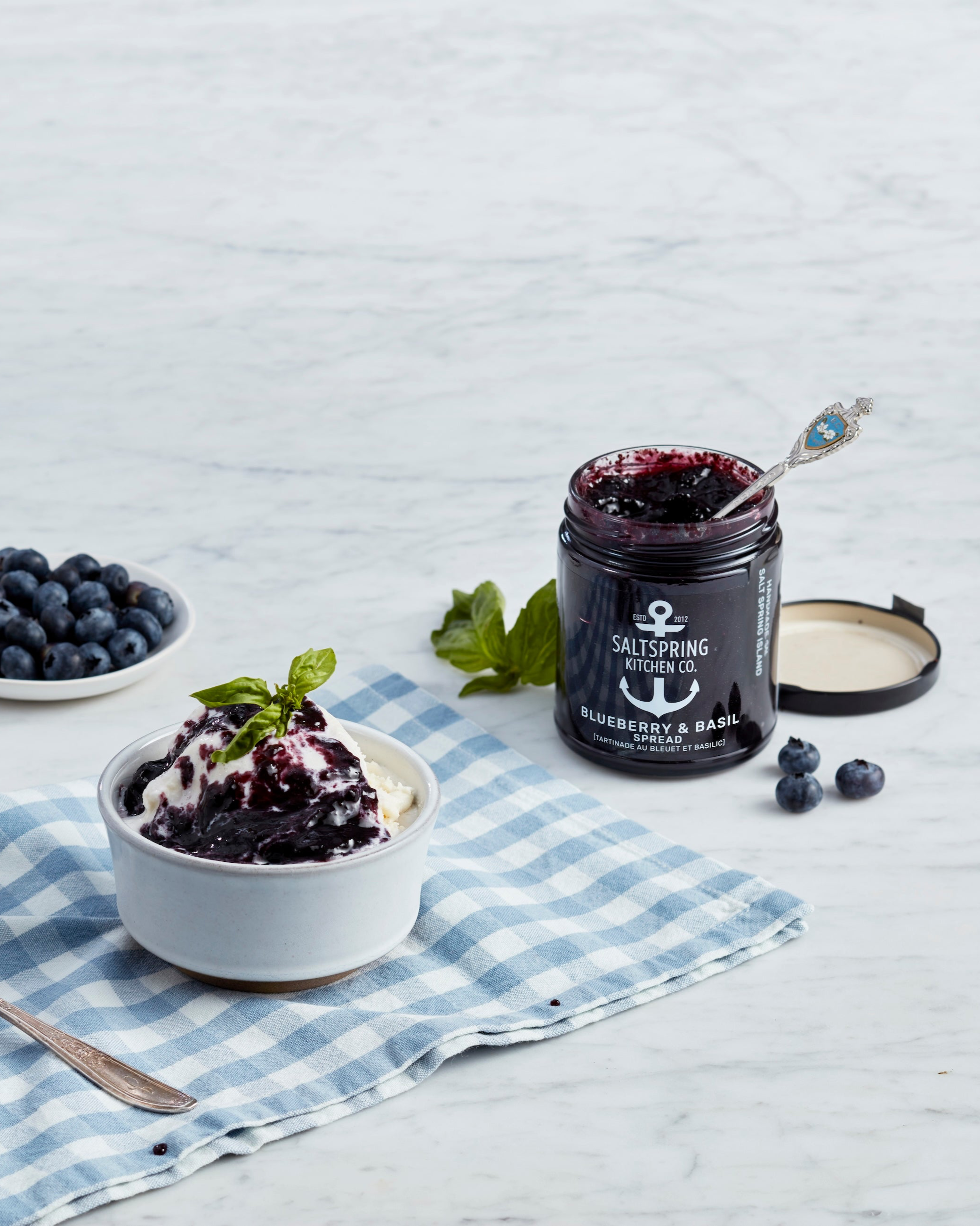 Jar of blueberry and basil jam with a bowl of dessert on a checkered cloth.