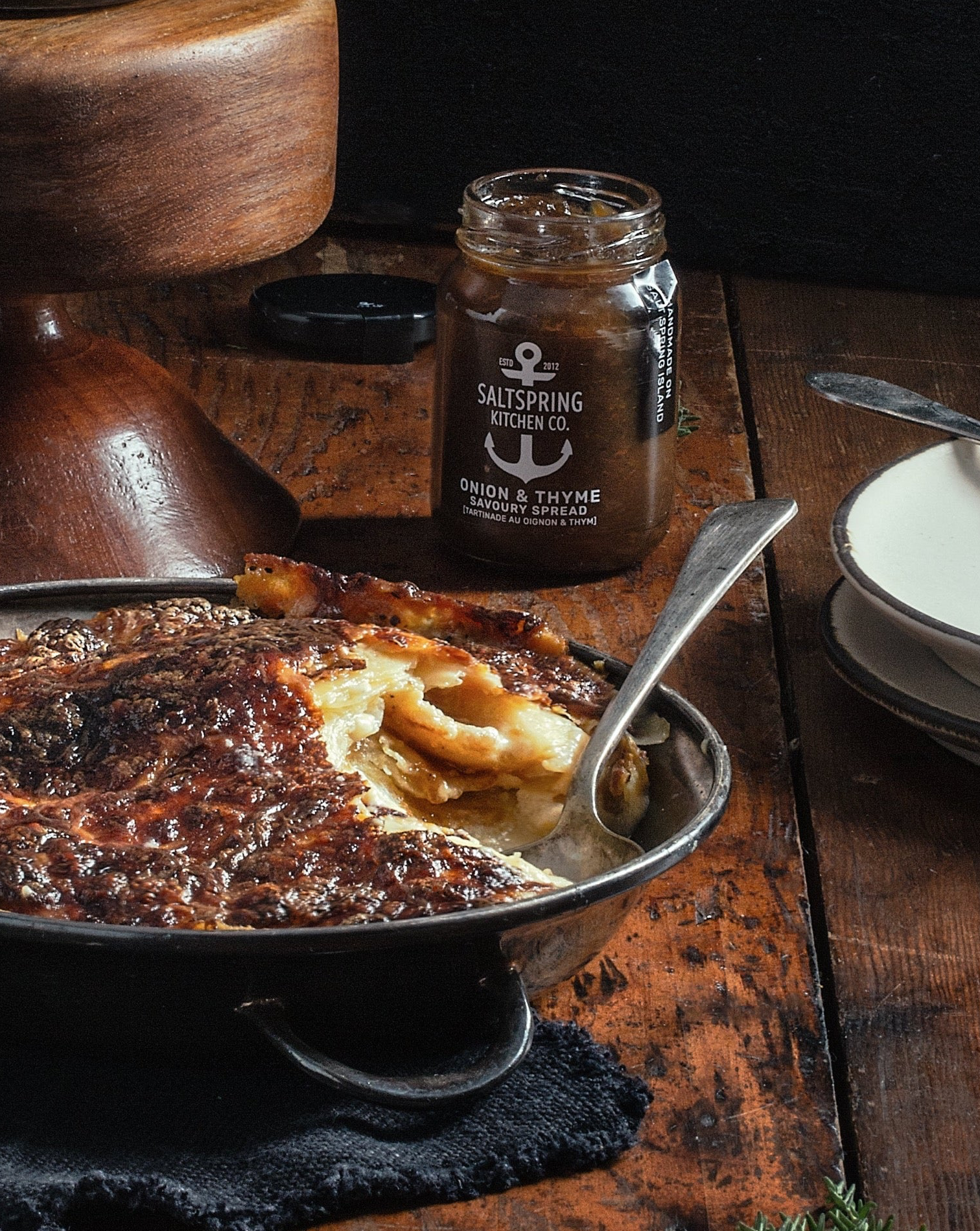 Baked dish of scalloped potatoes with a jar of SaltSpring Kitchen Co. Onion & Thyme Savoury Spread on a wooden table