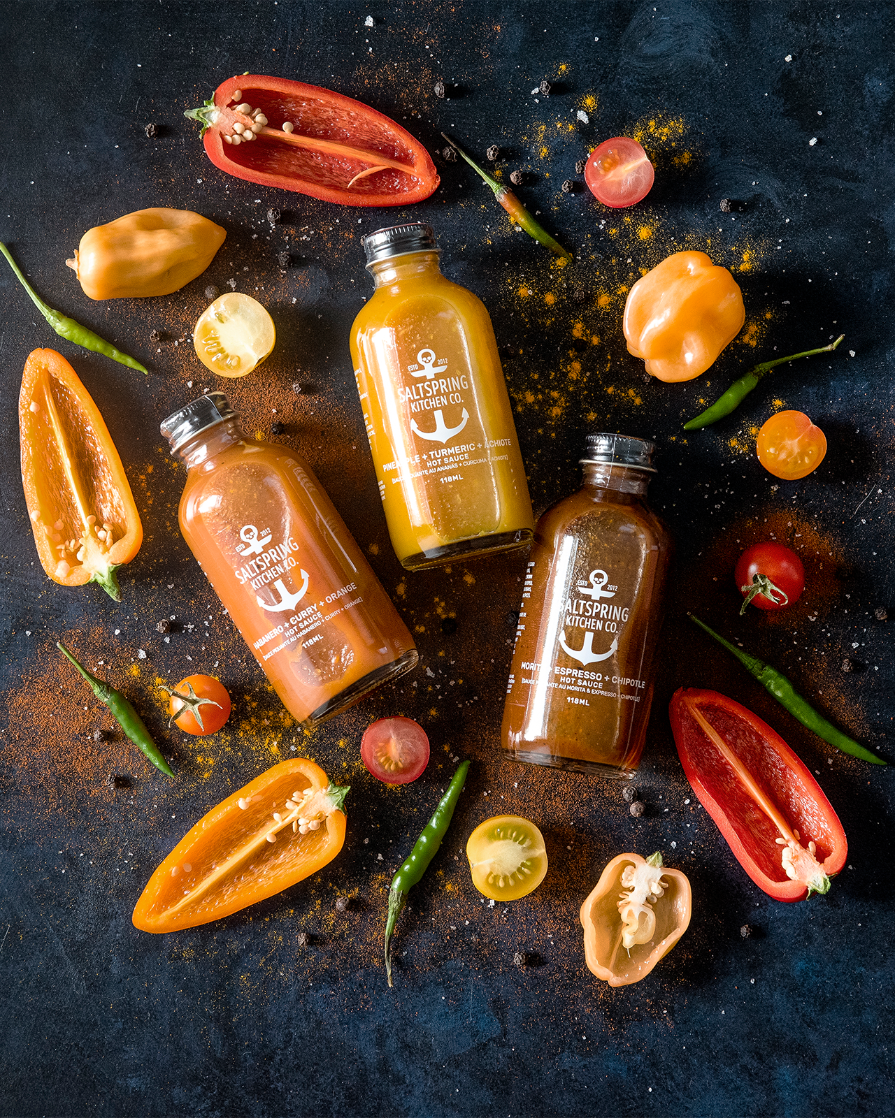 Three bottles of hot sauce with peppers and tomatoes on a dark surface
