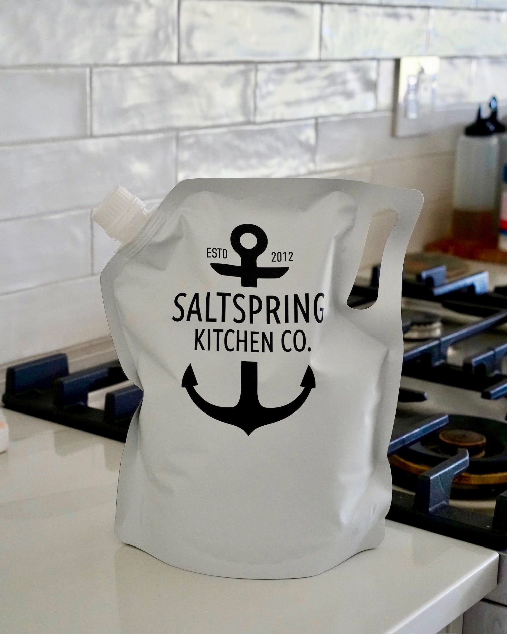 A white flexible refill pouch with SaltSpring Kitchen Company logo on it sitting on a kitchen counter in front of a tiled backsplash.