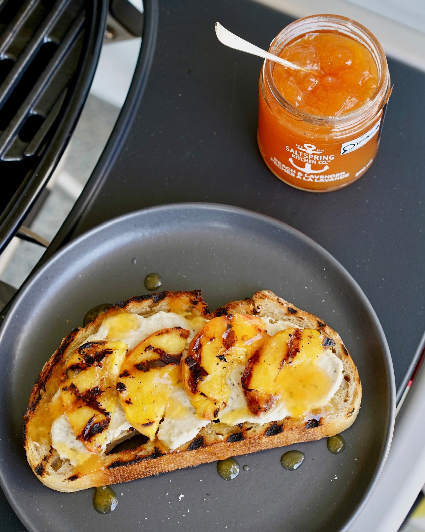 Grilled toast topped with soft cheese and grilled peaches on a dark plate, with a jar of Peach & Lavender Spread with a spoon next to the BBQ grill.