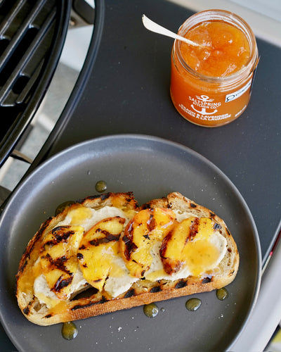 Grilled toast topped with soft cheese and grilled peaches on a dark plate, with a jar of Peach & Lavender Spread with a spoon next to the BBQ grill.