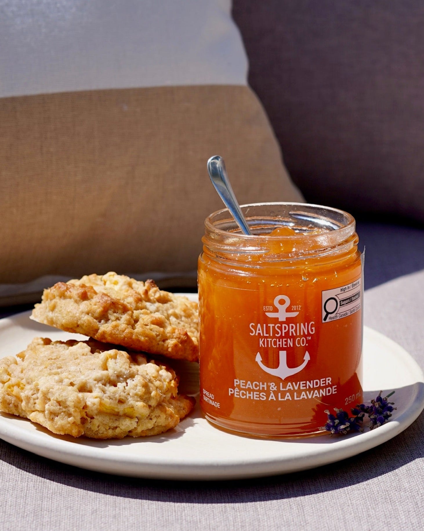 A jar of Saltspring Kitchen Co. Peach and Lavender Spread sits on a white plate with two peach scones, set on a light gray fabric loveseat surface with cushions in the background outside.