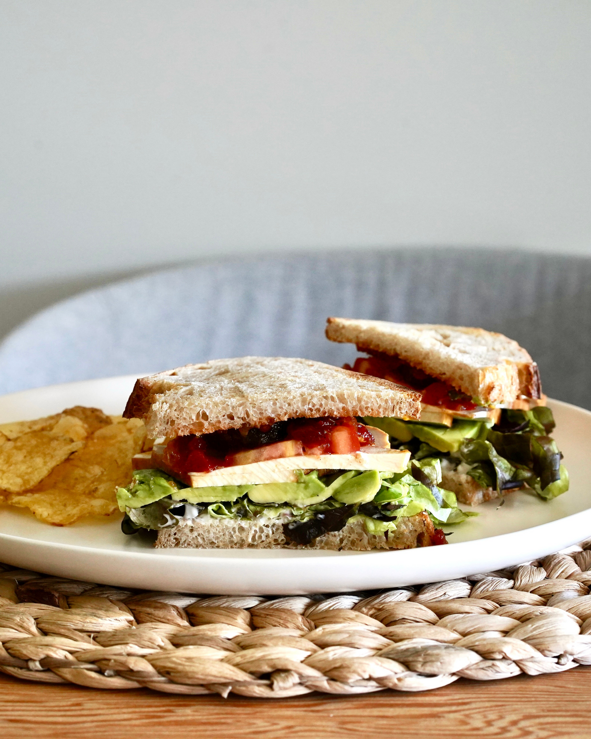 Vegetable sandwich cut in half on a plate with potato chips, placed on a woven mat.