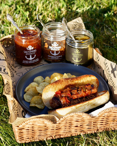 Vegan brat with SaltSpring Kitchen Company Smoky Red Relish and Charcuterie Mustard on a plate with potato chips and three jars of condiments in a picnic setting.