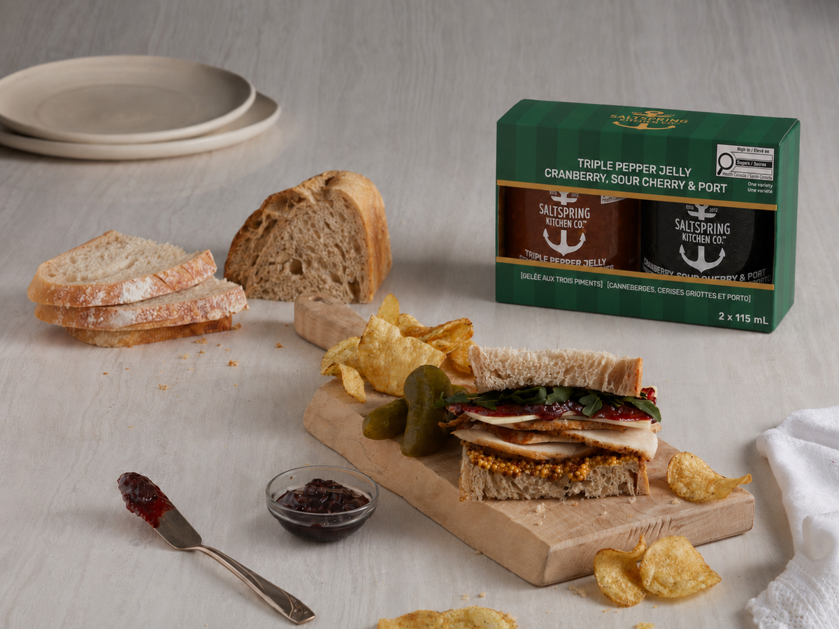 A Winter Duo Collection displayed on a tabletop with bread, chips, pickles, and a sandwich. The green box contains Triple Pepper Jelly, and Cranberry, Sour Cherry, and Port Preserves, with two jars visible through the front window.