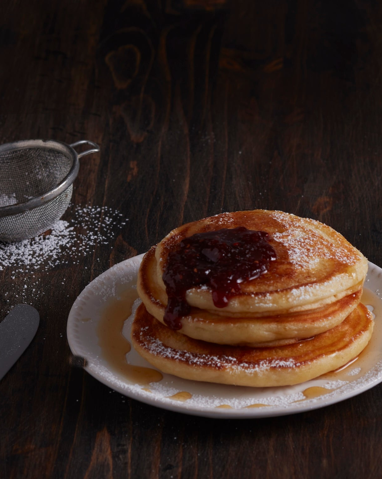 Stack of pancakes with syrup on a white plate with powdered sugar in the background