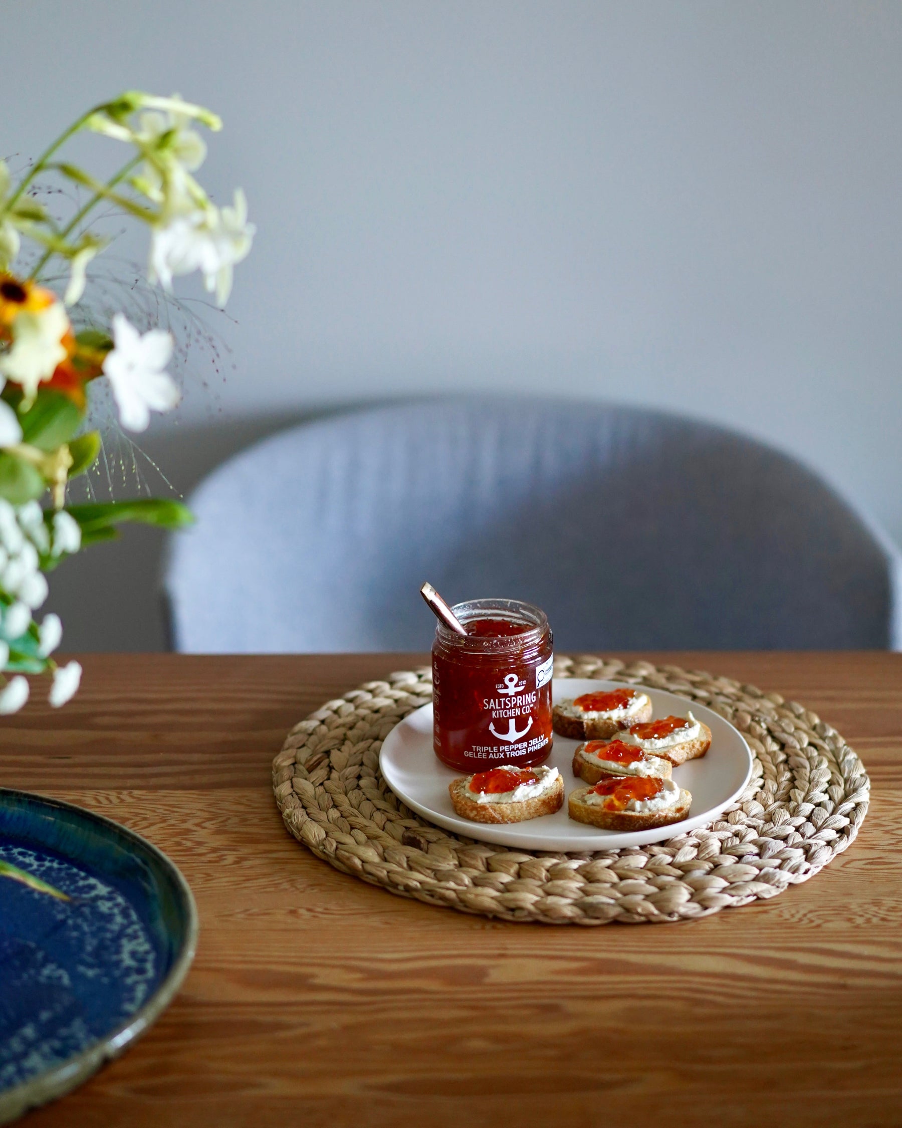 Jar of SaltSpring Kitchen Company Triple Pepper Jelly on a plate with crostini slices on a woven placemat, flowers in the background
